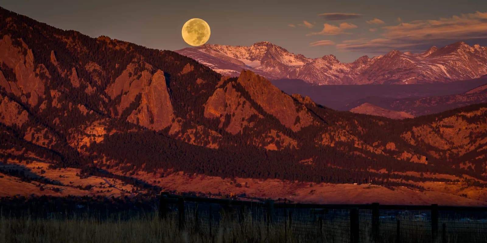 Moonrise over the Boulder Flatirons at dusk, with golden light illuminating tilted sandstone formations above forested slopes.